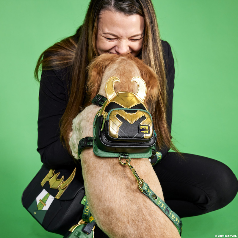 Woman wearing a dark suit and wearing the Loungefly President Loki crossbody bag smiling down at a dog with beige fur, who is facing her and wearing the Loki mini backpack dog harness. They both are against a green background. 